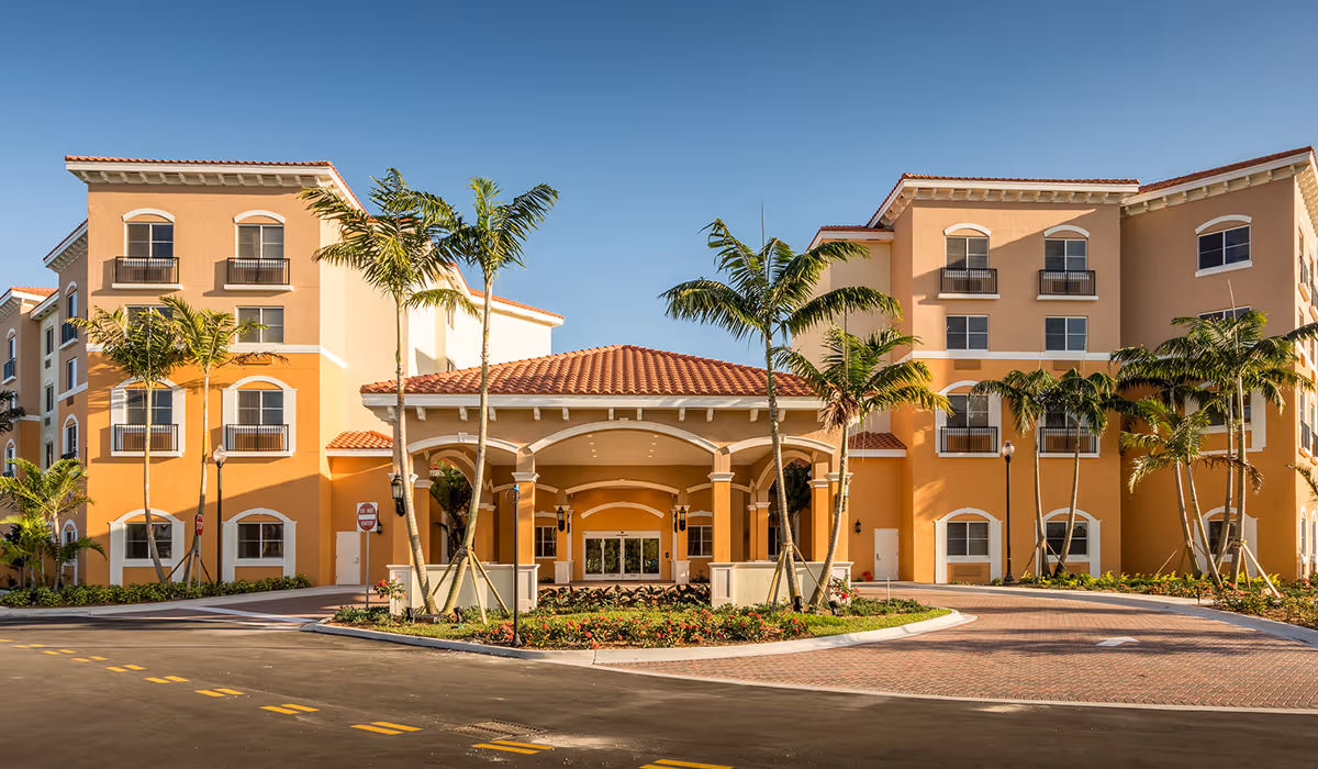 Front exterior view of The Palace Gardens - Assisted Living Retirement Community building with a covered entrance, palm trees, and a clear blue sky.