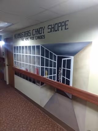 A hallway with a mural on the wall depicting a candy shop window and door. The mural includes the text 'Neimeisters Candy Shoppe Fine Home Made Candies'. There is a wooden handrail along the wall and patterned carpet on the floor.