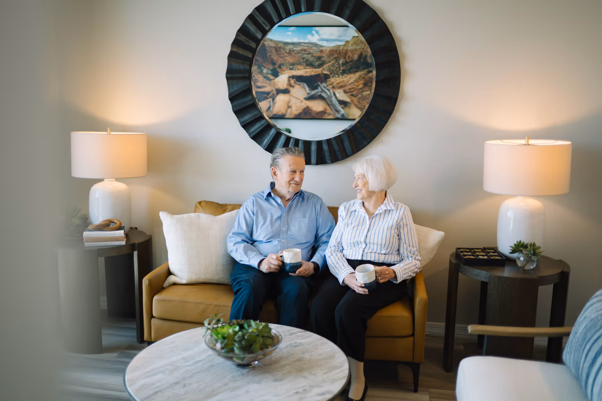 An elderly man and woman sitting on a mustard-colored couch in a cozy living room, each holding a coffee mug and smiling at each other. The room features two side tables with white lamps, a round marble coffee table with a small plant, and a large round mirror with a scenic landscape reflection on the wall behind them.
