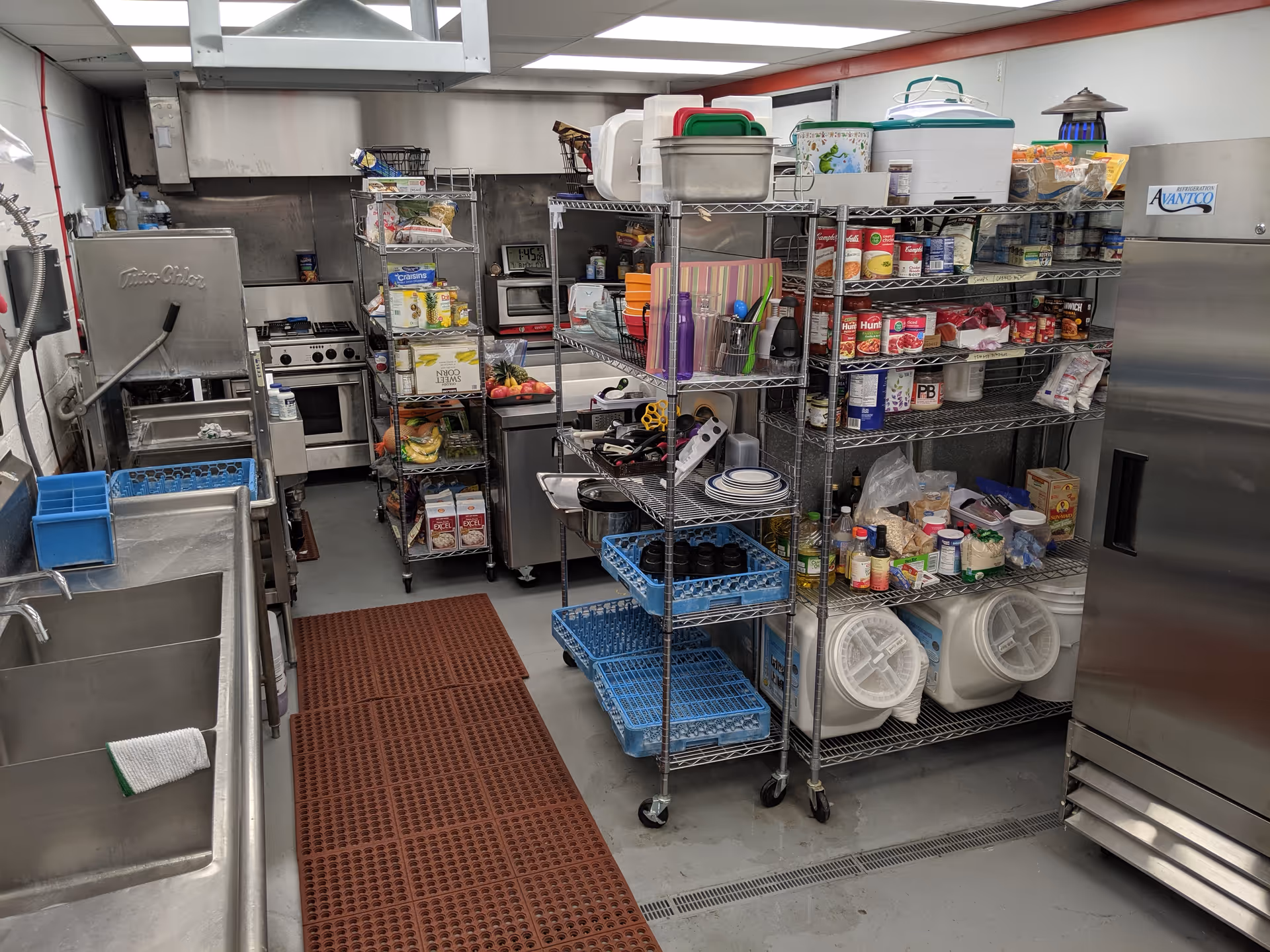 A commercial kitchen with stainless steel sinks on the left, metal shelving units stocked with food items and kitchen supplies in the center and right, a large refrigerator on the right, and a stove and microwave in the background.