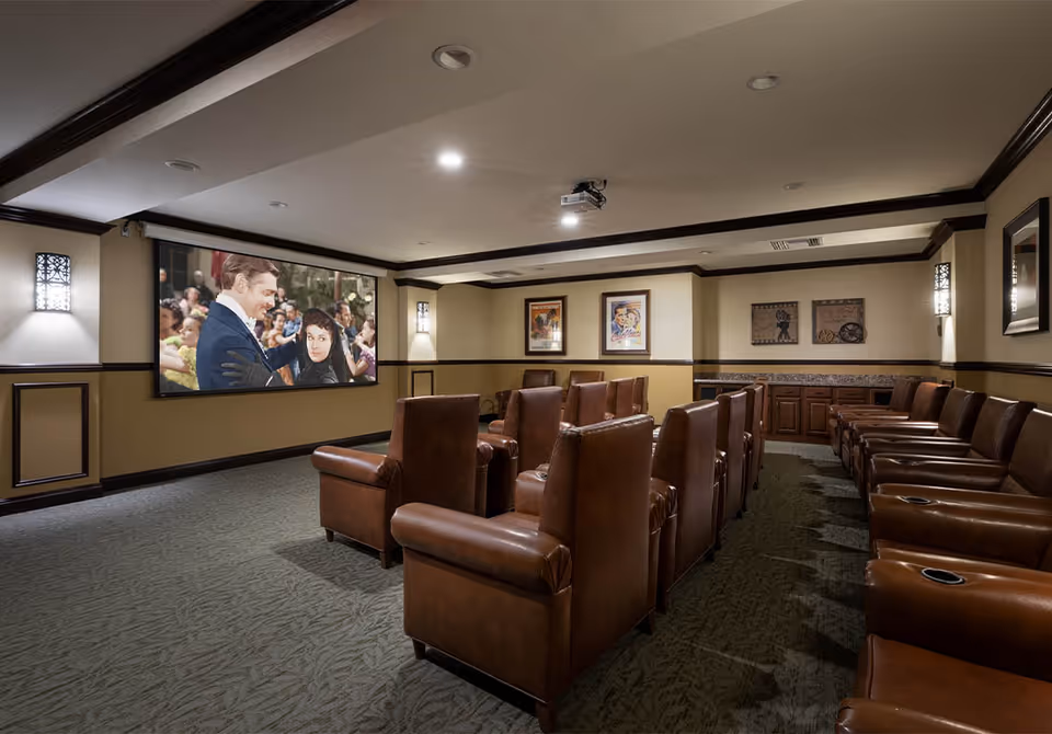 A cozy home theater room with two rows of brown leather recliner chairs facing a large projection screen displaying a classic movie scene. The room has beige walls with dark wood trim, framed movie posters on the walls, and soft lighting fixtures.