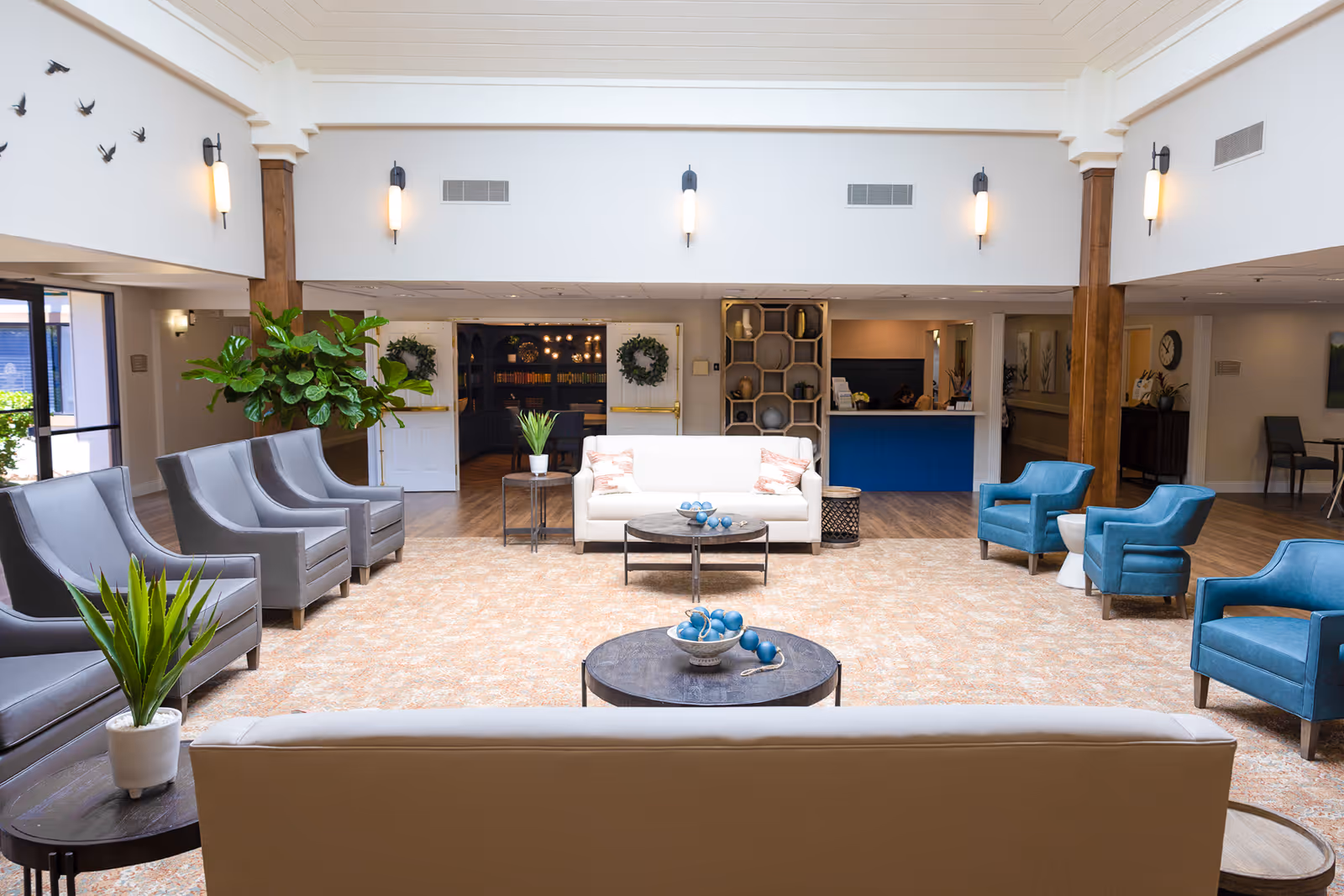 A spacious senior living facility common area with a beige sofa in the foreground, a white sofa with two pillows, several gray and blue armchairs arranged around circular coffee tables with decorative items, potted plants, and wall sconces. The room has a high ceiling with white beams and large windows letting in natural light. In the background, there is a reception desk with a blue front and an open doorway leading to another room with bookshelves and warm lighting.