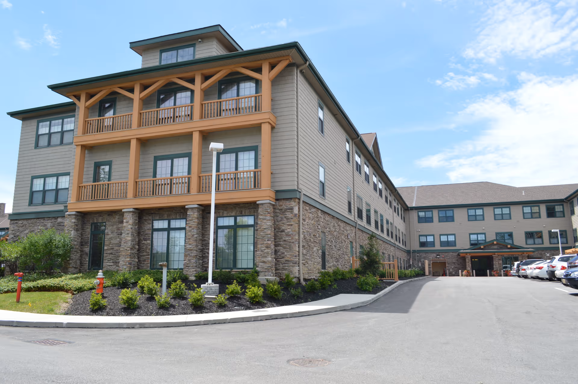 Exterior view of a multi-story senior living facility building with stone and wood siding, multiple windows, balconies, a parking area with cars, and a clear blue sky with some clouds.