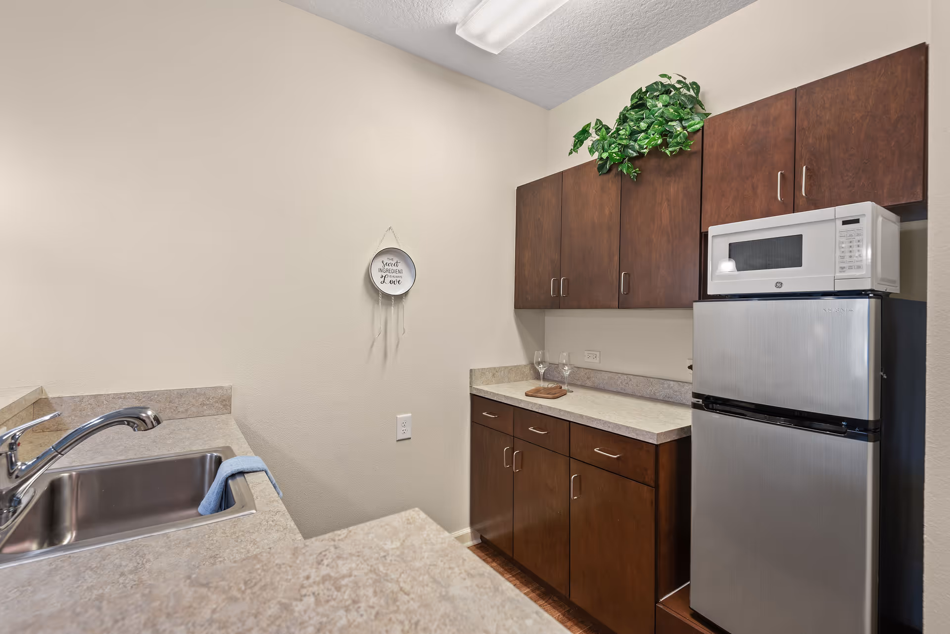 Small kitchen area with a stainless steel sink and faucet on the left, beige countertops, dark wooden cabinets above and below the counter, a small silver refrigerator with a white microwave on top, two wine glasses and a small cutting board on the counter, and a green plant on top of the upper cabinets. A decorative plate hangs on the wall with the text 'The secret ingredient is always love.'