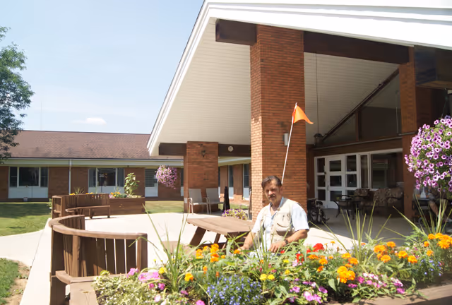 A man sitting on a bench in a garden area outside a brick building with a covered entrance. The garden has colorful flowers and greenery, and the building has large windows and a sloped roof.
