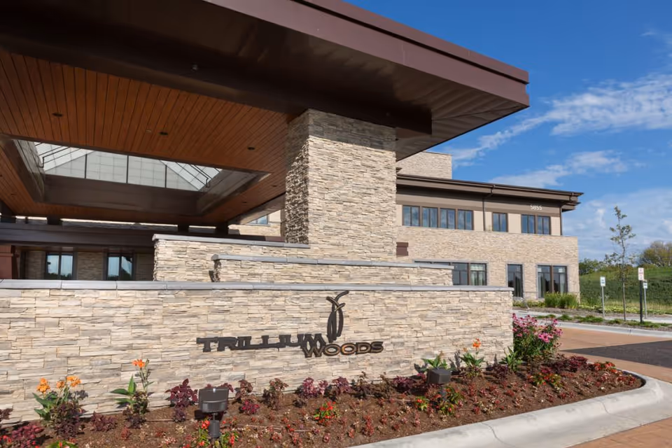 Exterior view of the Trillium Woods senior living facility showing a modern stone facade with the facility name displayed on a stone wall surrounded by landscaped flowers and plants under a blue sky with some clouds.
