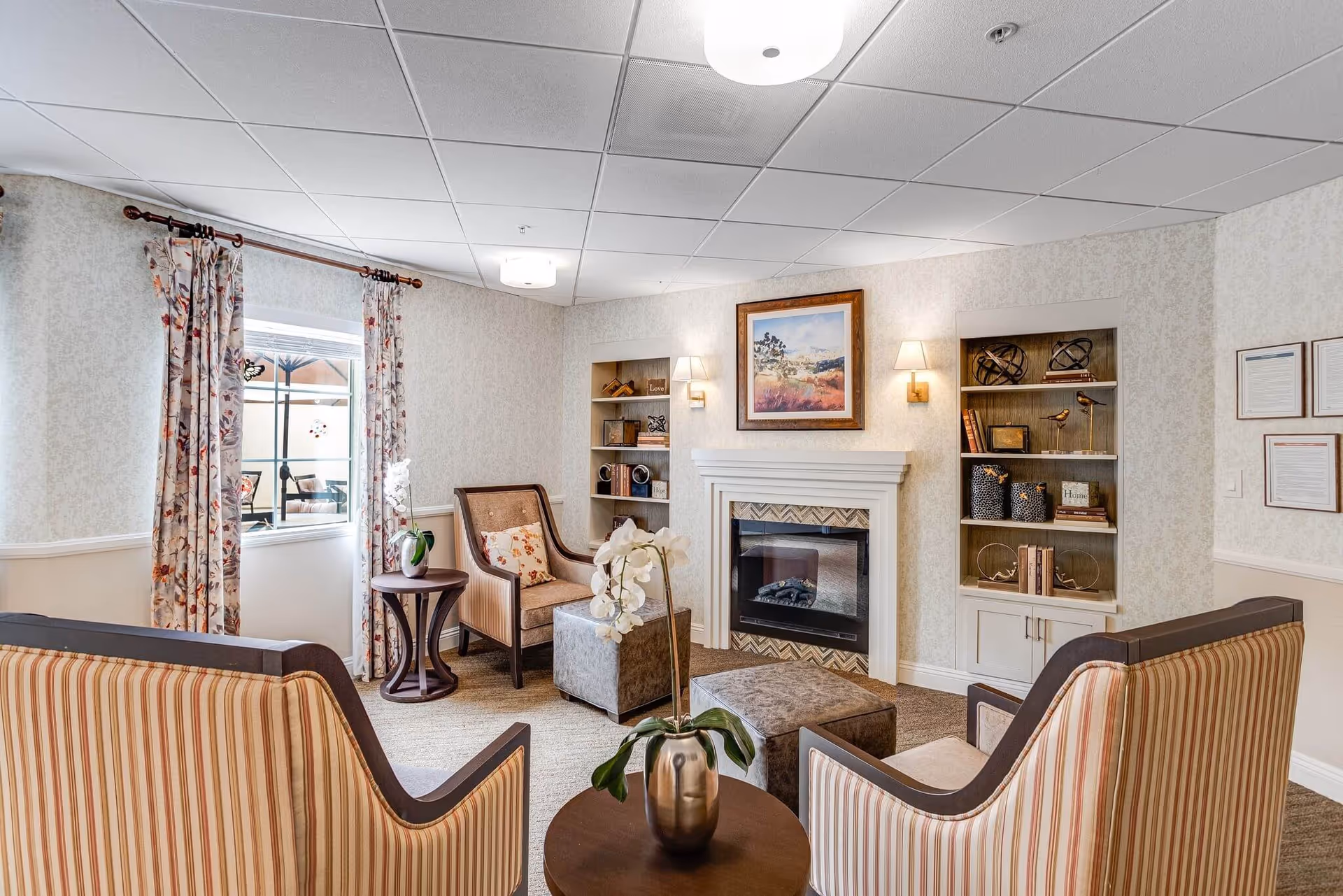 A cozy living room area in a senior living facility with two striped armchairs, a cushioned armchair, two ottomans, a small round table with a vase of white flowers, a fireplace with a framed landscape painting above it, built-in shelves with decorative items, and floral curtains on the window.