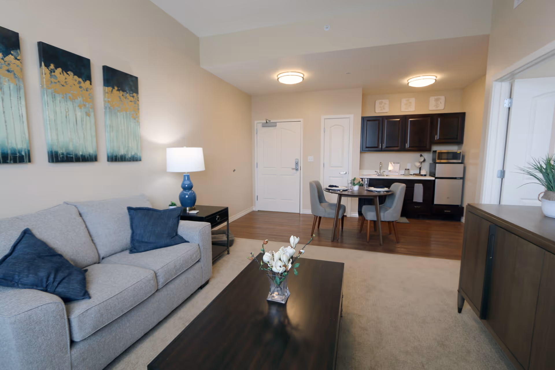 A cozy living area in an assisted living facility featuring a gray sofa with dark blue pillows, a dark wooden coffee table with a vase of white flowers, a side table with a blue lamp, and three abstract paintings on the wall. In the background, there is a small dining table with four chairs and a kitchenette with dark cabinets, a microwave, and a mini fridge.