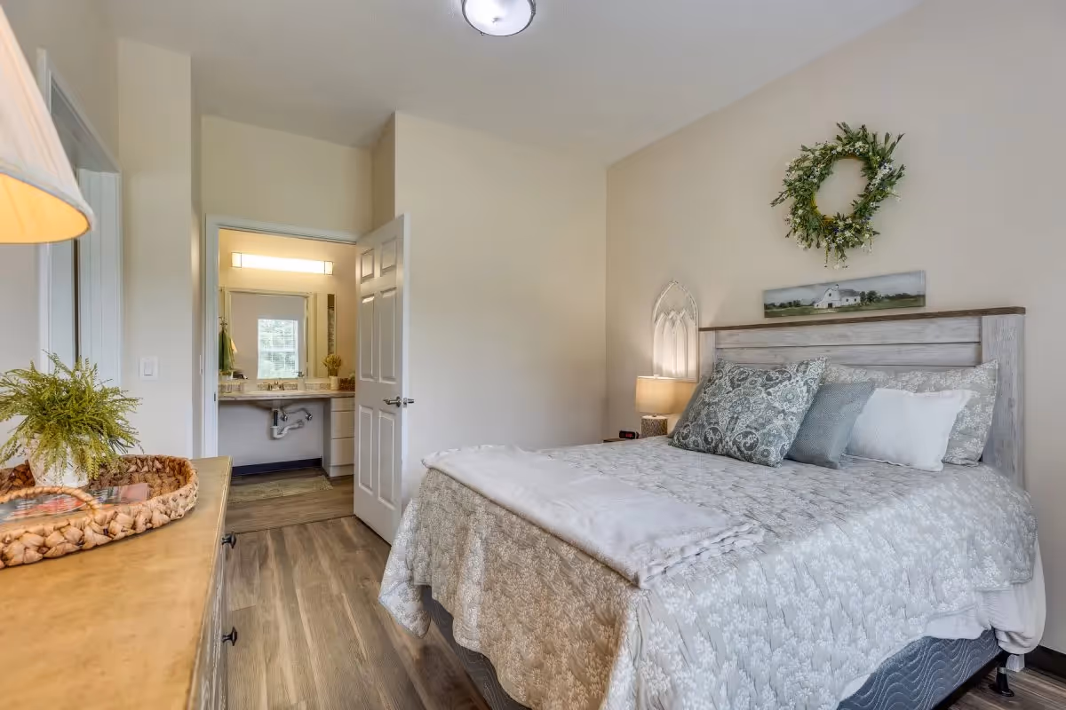 A cozy bedroom with a neatly made bed featuring patterned bedding and multiple pillows. A bedside table with a lamp and decorative wall art including a wreath and a landscape painting are visible. The room has wooden flooring and a dresser with a plant on top. An open door reveals a bathroom with a sink and mirror.