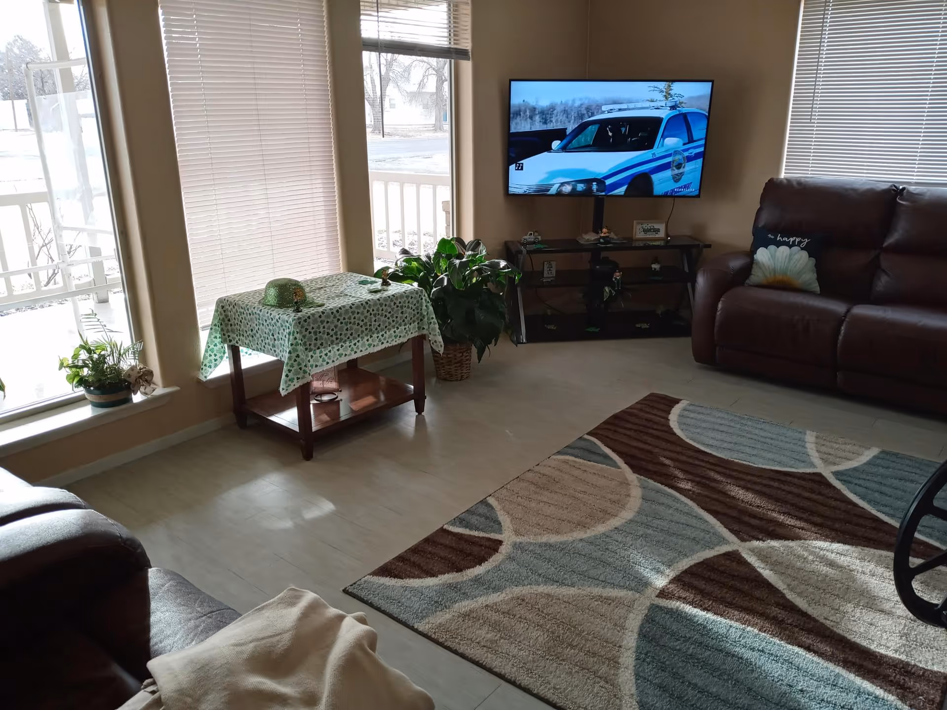 A cozy living room with large windows covered by blinds, a small table with a green patterned tablecloth and a green hat on top, a potted plant beside the table, a flat-screen TV mounted on the wall showing a police car, a brown leather sofa with a decorative pillow, and a patterned area rug on the floor.