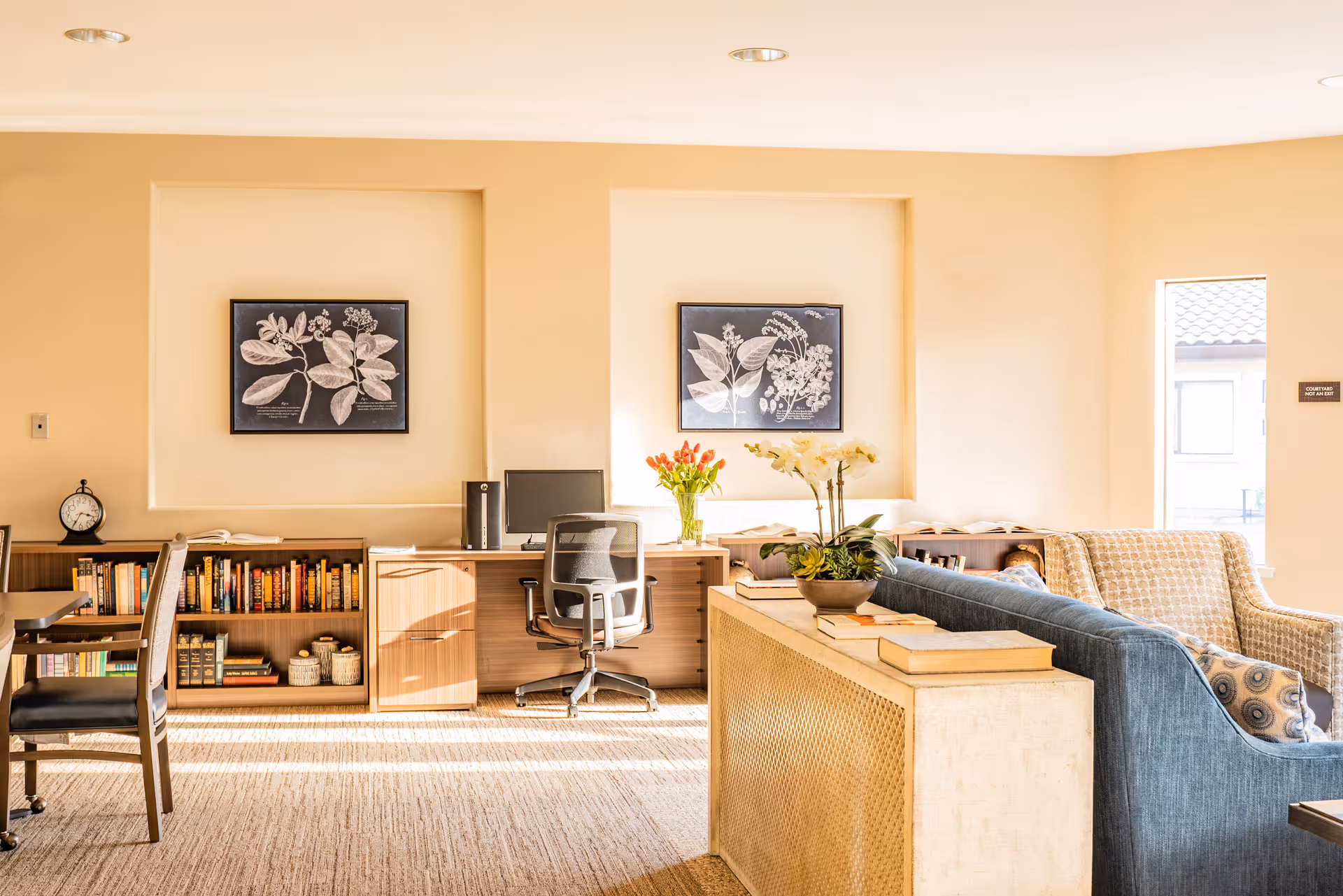 A bright and cozy interior room with beige walls and carpeted floor. The room features a wooden desk with a computer and office chair, bookshelves filled with books, two framed botanical prints on the wall, a blue sofa with patterned cushions, and a small table with flowers and books. Sunlight streams in through a window on the right side.