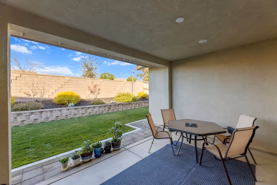 Covered backyard patio with a table and four chairs, potted plants along the edge, and a grassy yard with a low retaining wall under a blue sky.