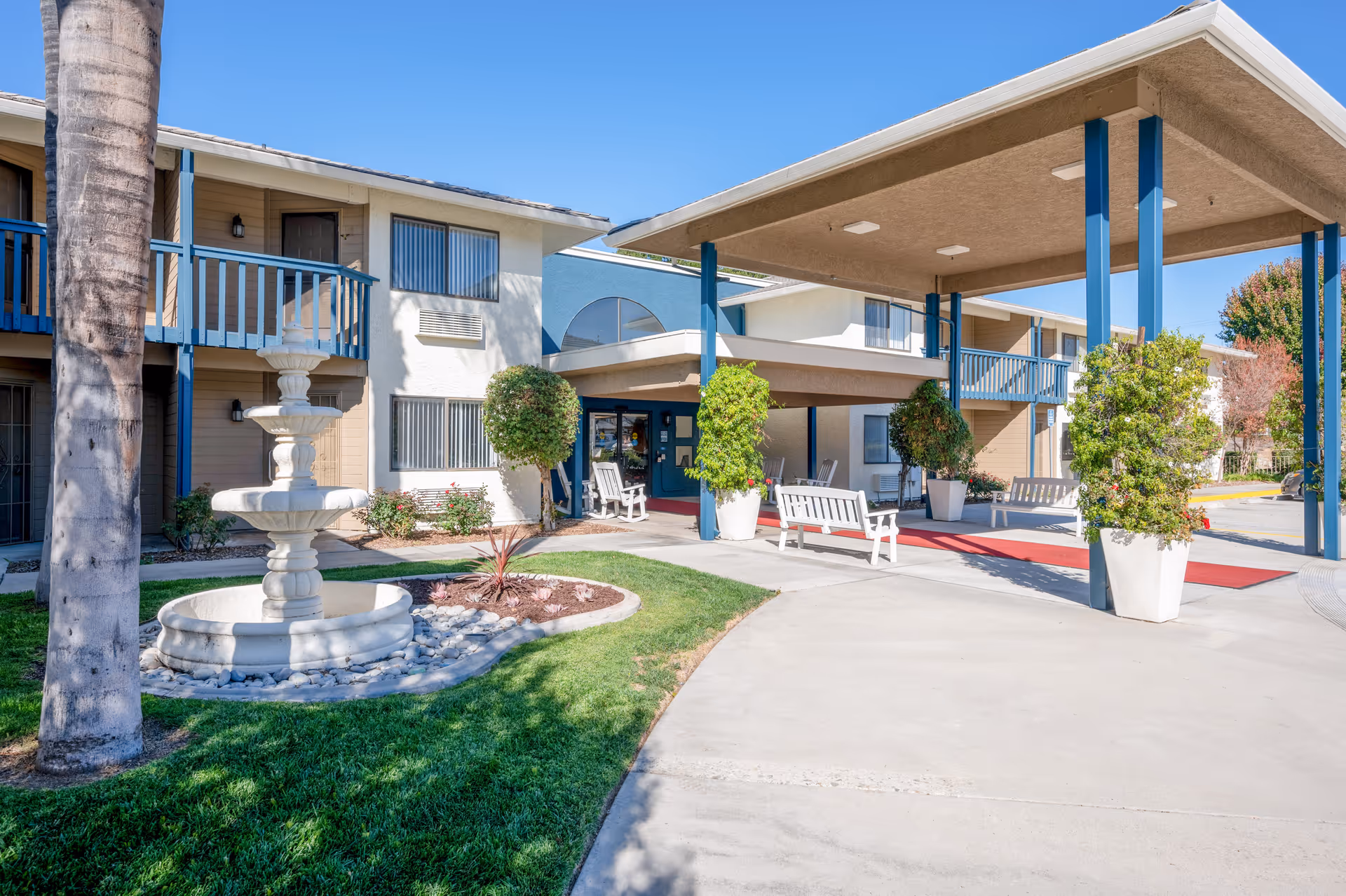 Exterior view of Truewood by Merrill, West Covina senior living facility showing a covered entrance with blue support beams, white benches, potted plants, a three-tiered white fountain, and a two-story building with balconies and windows under a clear blue sky.