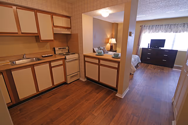 Interior view of a senior living facility apartment showing a kitchen area with wooden cabinets, a sink, and a stove on the left. Adjacent to the kitchen is a small counter with two place settings. Beyond the counter is a bedroom area with a bed, a bedside lamp, a dresser, and a television near a window with curtains. The floor is wooden throughout.