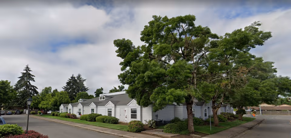 Low-rise white senior living buildings along a tree-lined street with manicured shrubs and a sidewalk.