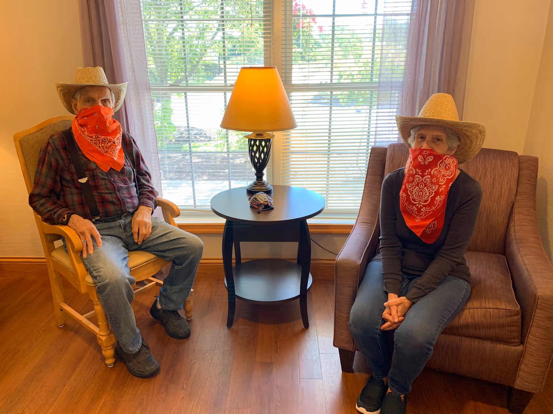 An elderly man and woman sitting indoors in front of a large window with blinds. Both are wearing straw cowboy hats and red bandanas covering their faces. The man is seated in a wooden armchair with a plaid shirt and jeans, while the woman is seated in a cushioned armchair wearing a dark long-sleeve shirt and jeans. A round side table with a lamp is positioned between them.