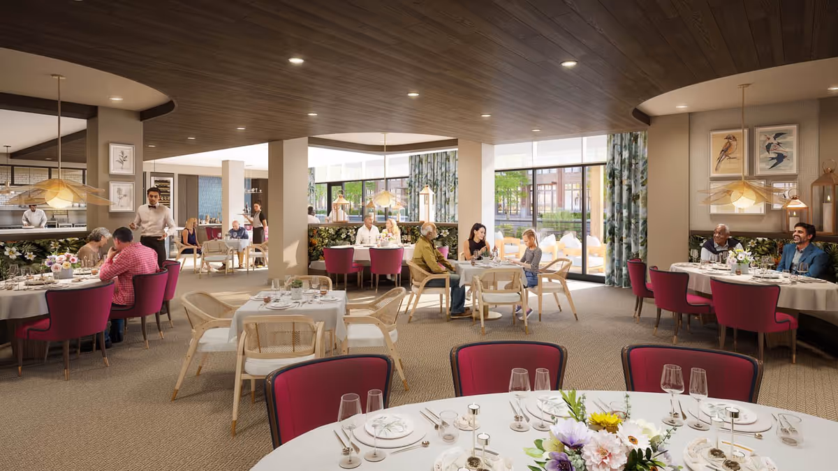 A bright and spacious dining room in a senior living facility with multiple tables set for meals. Several groups of people are seated and engaged in conversation while waitstaff attend to them. The room features large windows with floral curtains, modern light fixtures, and a warm, inviting atmosphere with a mix of purple and beige chairs.