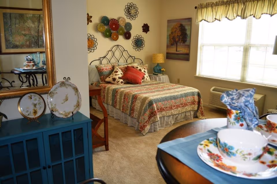A cozy bedroom with a bed covered in a colorful striped quilt and multiple pillows. The wall above the bed is decorated with metal floral art. To the right of the bed is a window with a valance curtain and an air conditioning unit below it. A small table with a lamp and a painting of a tree is next to the bed. In the foreground, there is a round table set with floral-patterned dishes and a blue napkin. On the left side, a blue cabinet holds decorative plates and a large mirror reflecting part of the room.