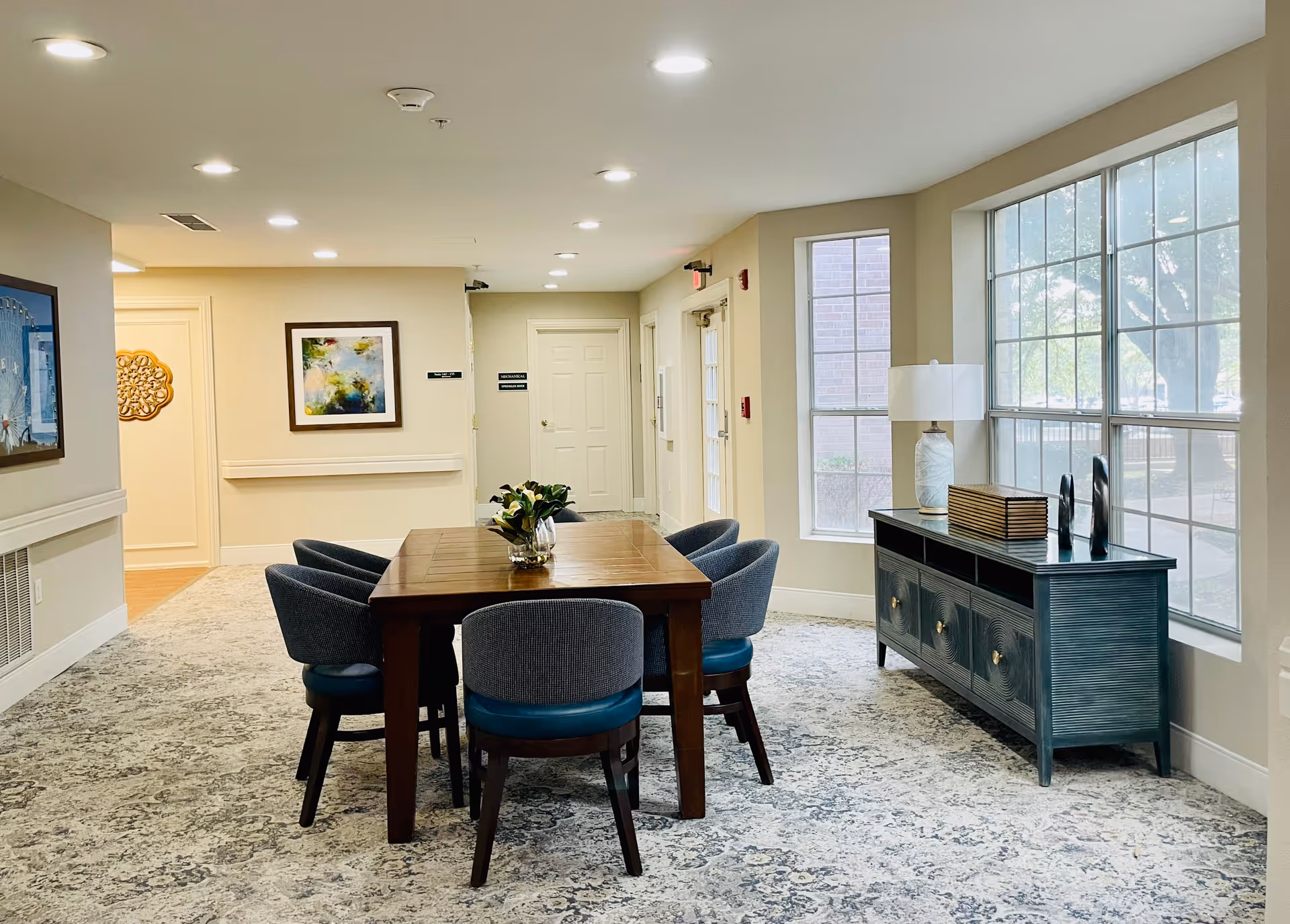 Well-lit communal dining area with a wooden table surrounded by upholstered chairs, a sideboard under large windows, and artwork on the walls.