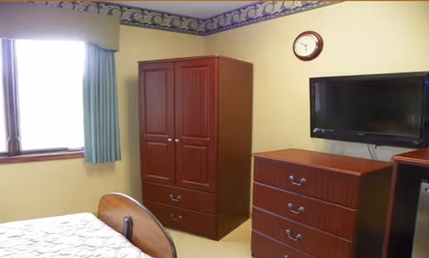 A simple bedroom with a wooden bed, wardrobe, dresser, wall-mounted TV, clock, and a curtained window.