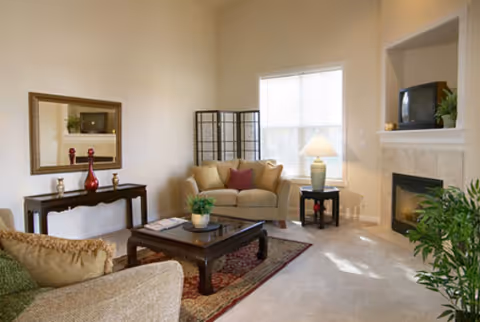 A cozy living room with beige walls and carpet, featuring a beige loveseat with a red pillow, a beige armchair, a dark wooden coffee table with a plant and magazines, a side table with a lamp, a fireplace with a TV on the mantel, a decorative folding screen, a console table with red vases, and a large window letting in natural light.