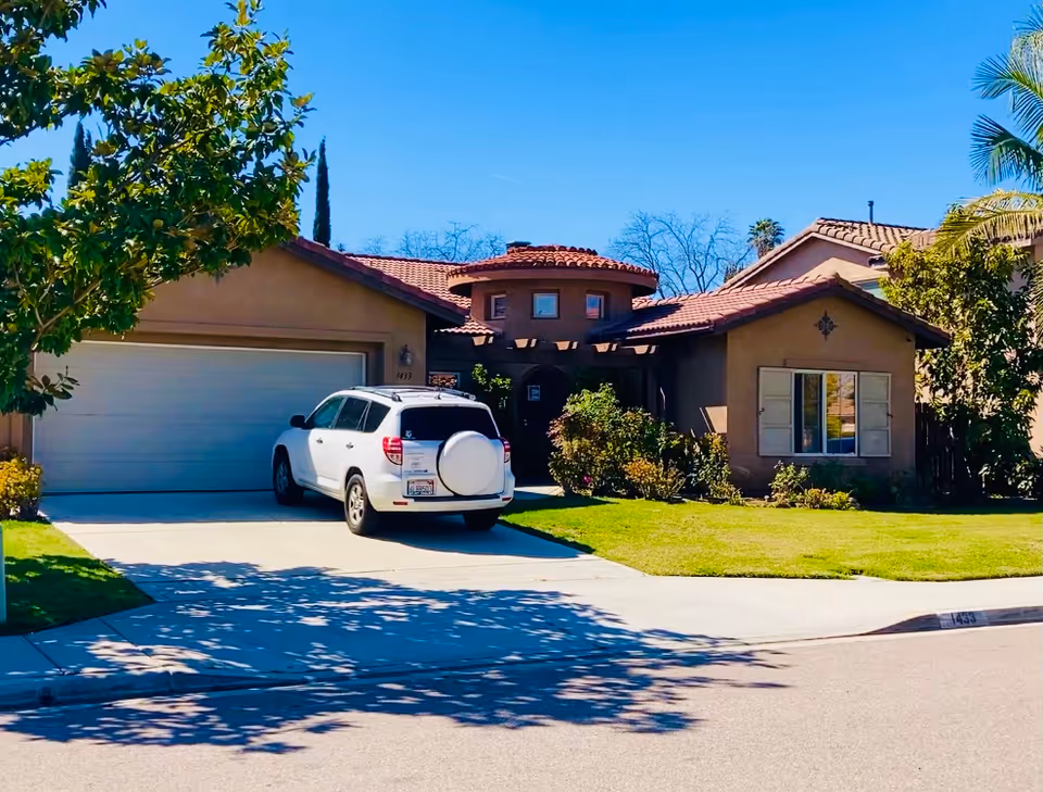 Front exterior view of a single-story residential building with a tiled roof, a white SUV parked in the driveway, green lawn, and trees under a clear blue sky.