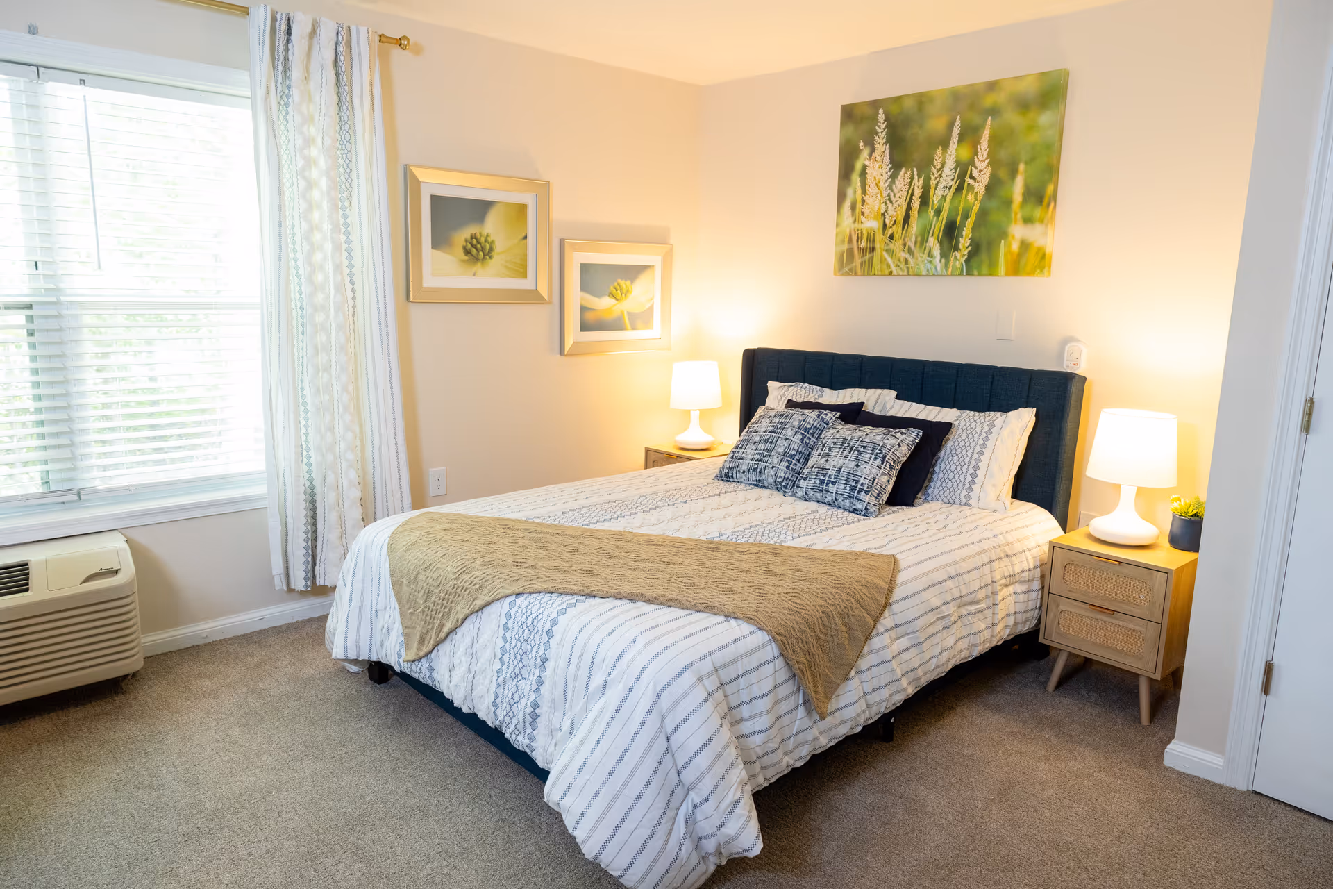 A bright furnished bedroom with a made bed, two nightstands with lamps, a window with curtains, and wall art above the headboard.