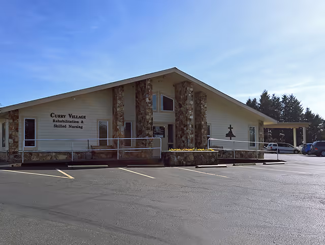 Front exterior of the Curry Village Rehabilitation & Skilled Nursing building with stone columns, an entry ramp, and an empty parking lot under a clear sky.