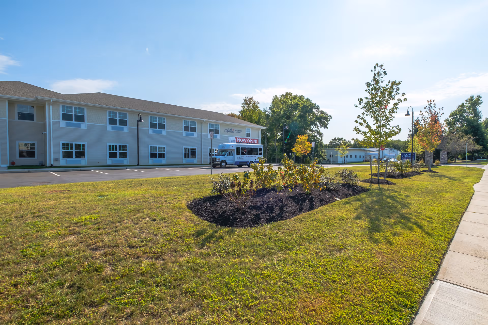Exterior view of All American Assisted Living at Tinton Falls building with a parking lot, a shuttle bus parked near the entrance, landscaped garden beds with small trees and shrubs, and a sidewalk on a sunny day.