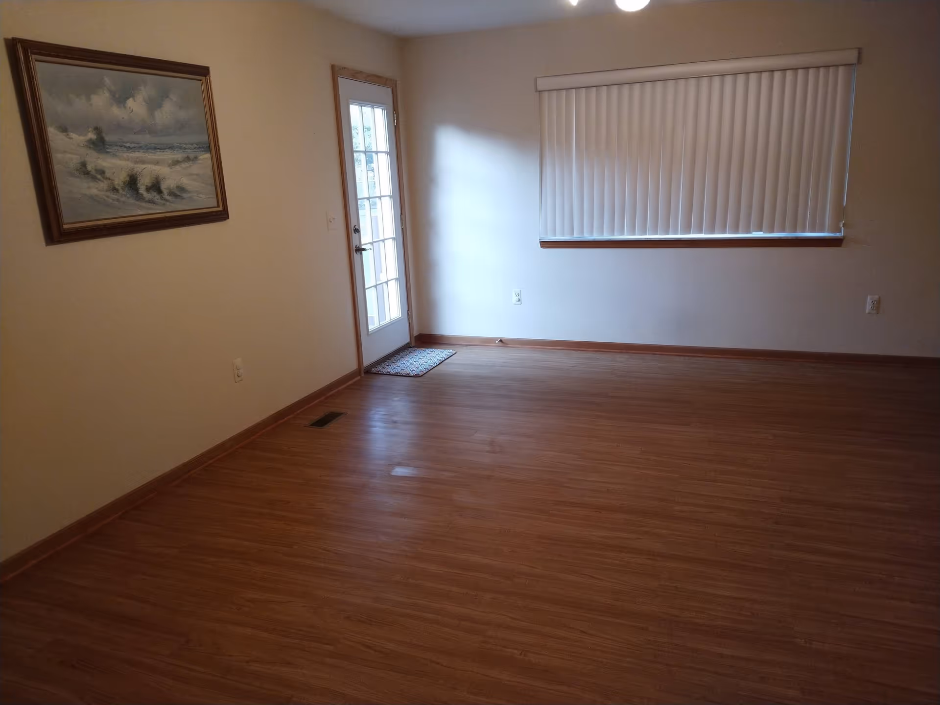 Empty living room with wood floors, a glass door, vertical blinds, and a framed seascape painting on the wall.