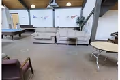 Interior view of a senior living facility common area with beige sofas, a pool table, folding tables, and chairs. The room has a high ceiling with exposed wooden beams and white walls decorated with pennants.