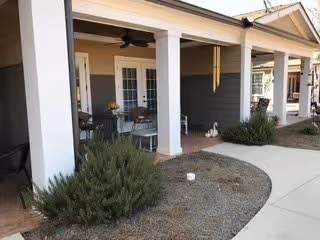 Outdoor covered patio area with white columns and ceiling fans, featuring seating with tables and chairs. There are shrubs and a curved concrete walkway in front of the patio, part of a residential or senior living facility exterior.