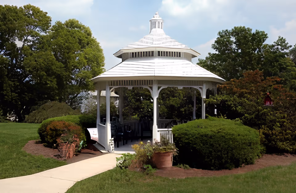 A white gazebo with a tiered roof situated in a well-maintained garden area with green bushes, trees, and a paved walkway leading to the gazebo.