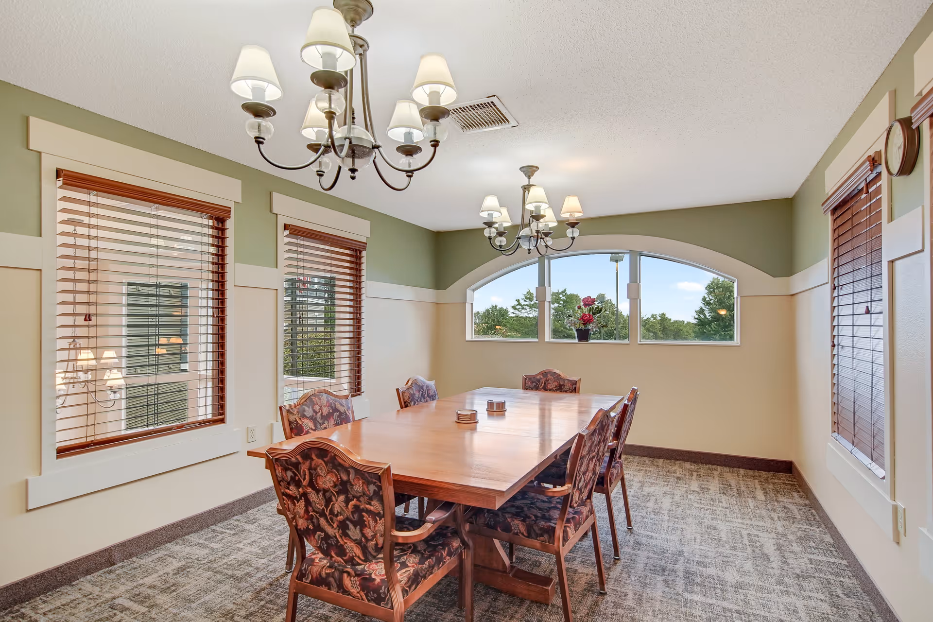 A dining room with a wooden table and six upholstered chairs with floral patterns. The room has three windows with wooden blinds, two chandeliers with lampshades hanging from the ceiling, and a small plant on the windowsill. The walls are painted in light green and beige tones, and the floor is carpeted.