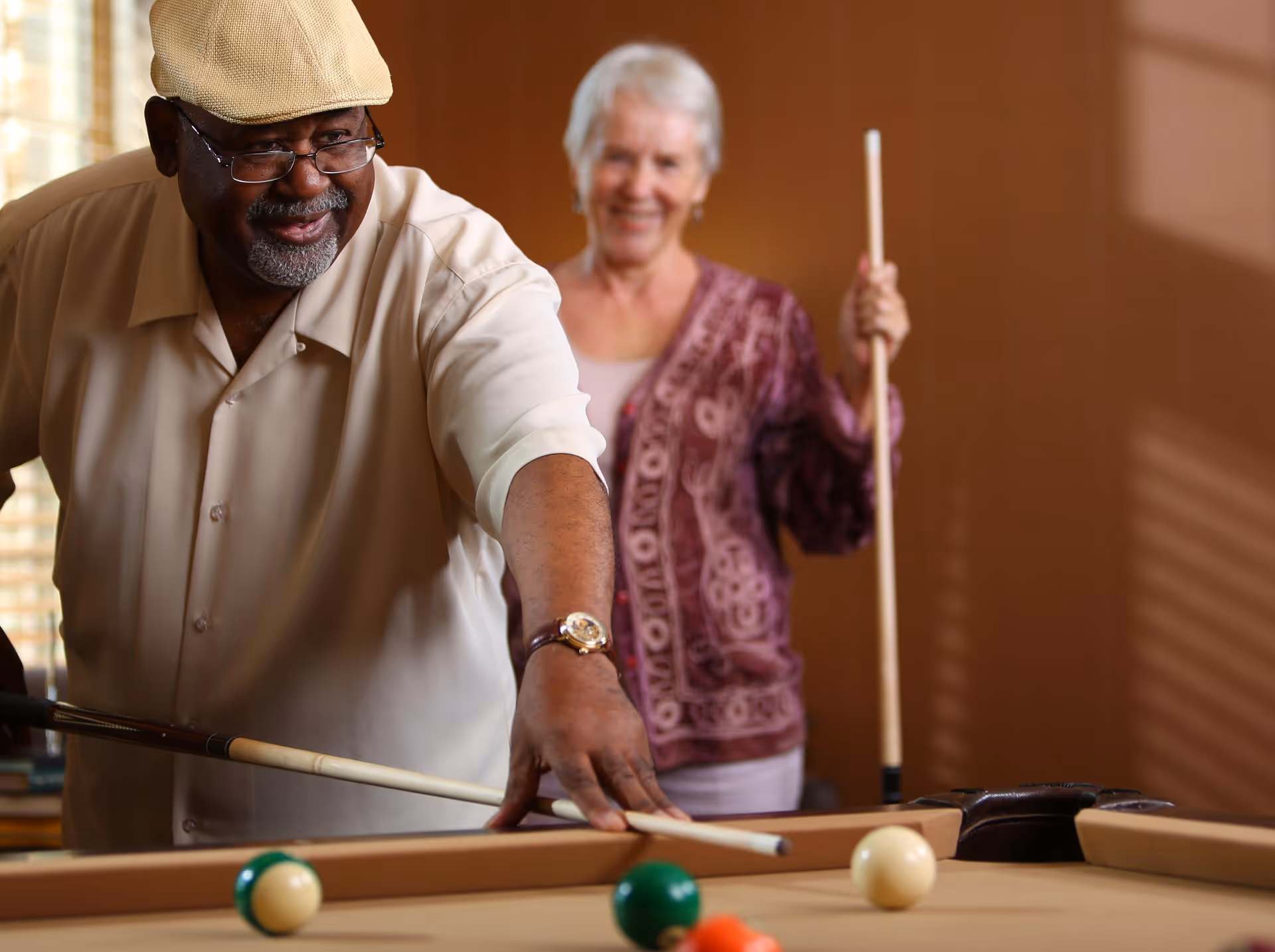 An elderly man and woman playing pool indoors. The man is aiming with a pool cue while the woman stands behind him smiling, holding her own cue stick.