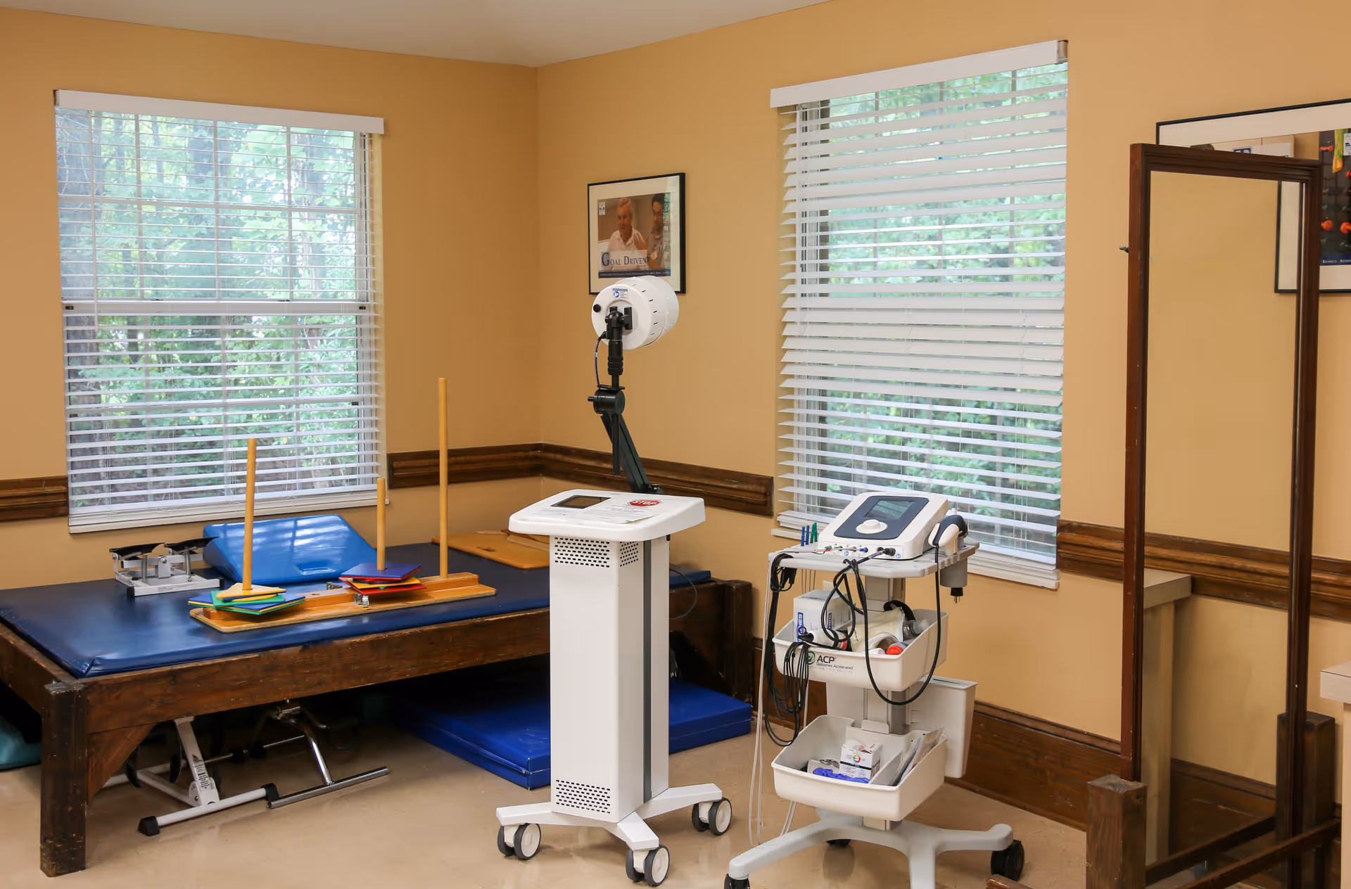 A rehabilitation therapy room with a padded therapy table holding various therapy tools, including wooden rings and blocks. Two windows with white blinds let in natural light. There is medical equipment on wheeled carts, including a device with cables and a light therapy machine. The walls are beige with dark wood trim, and a large standing mirror is visible on the right side.