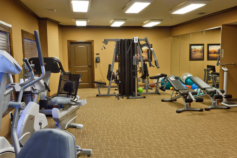 Indoor fitness room with exercise equipment including stationary bikes, weight machines, benches, and exercise balls. The room has beige walls, carpeted floor, a large mirror on one wall, and framed artwork.