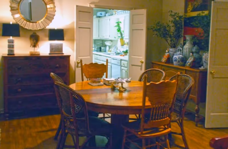 A cozy dining room with a round wooden table surrounded by six chairs, two wooden and four wicker. The room features wooden flooring, a dark wooden chest of drawers with two lamps and a decorative mirror above it on the left side, and a sideboard with various blue and white ceramic vases and figurines on the right. In the background, double doors open to reveal a white kitchen with cabinets, a sink, and some plants.