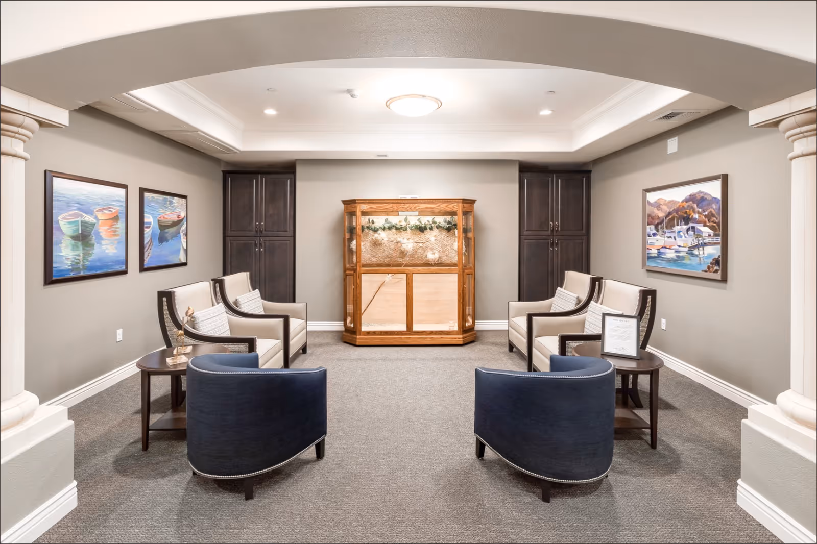 A cozy seating area in a memory care facility with two blue armchairs and four beige armchairs arranged around two small round wooden tables. The room has neutral-colored walls with three framed paintings of boats on the water. There are two dark wooden cabinets flanking a wooden display cabinet with glass doors in the center. The space is well-lit with a ceiling light and recessed lighting.