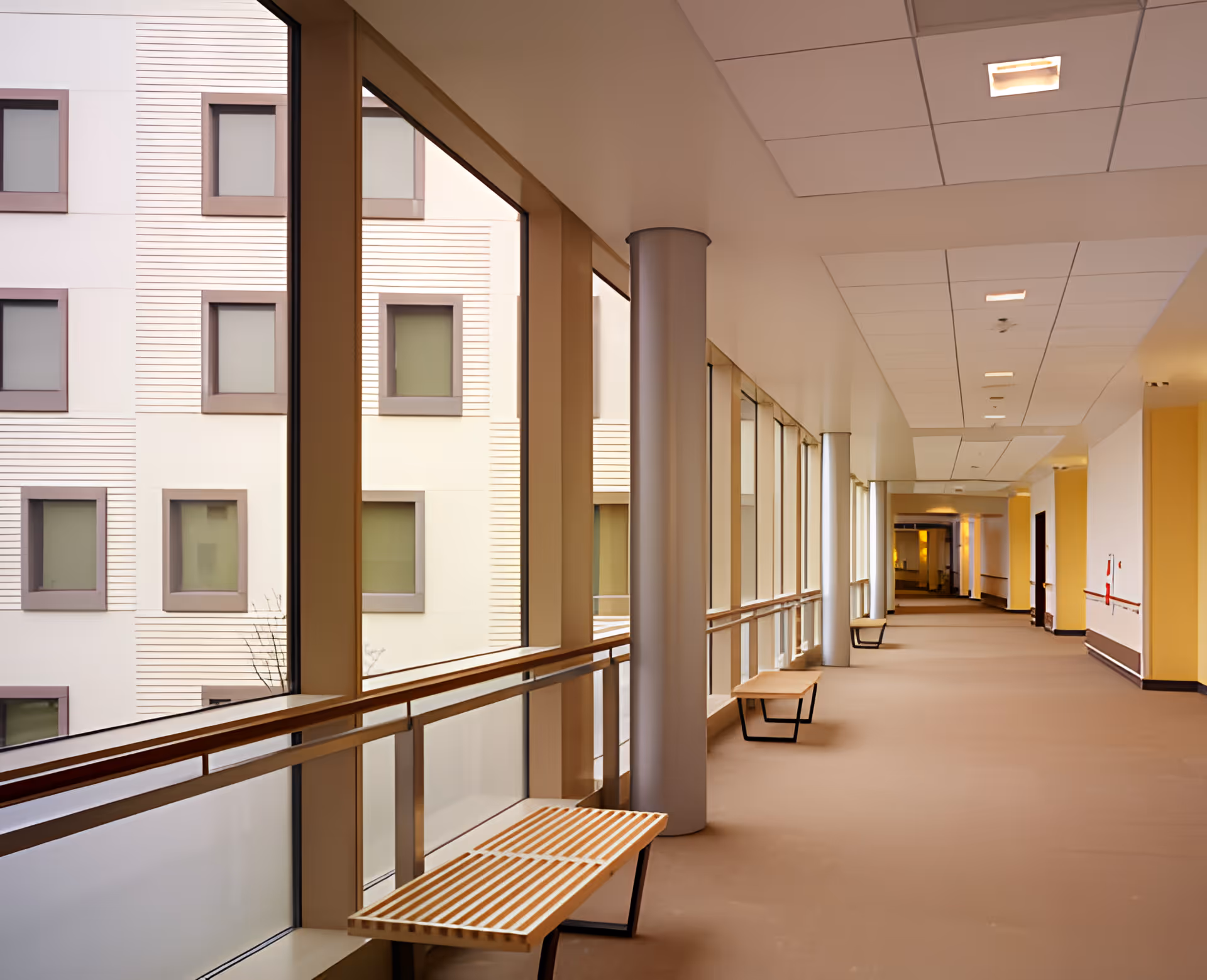 Bright, wide indoor corridor with large windows, benches, handrails and views of adjacent building windows.