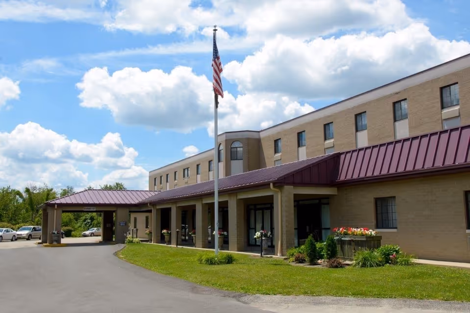 Exterior view of Southwestern Nursing and Rehabilitation Center building with a beige brick facade and a maroon metal roof. There is a flagpole with an American flag in front of the building, a driveway leading to the entrance, and several parked cars. The sky is partly cloudy with blue patches.