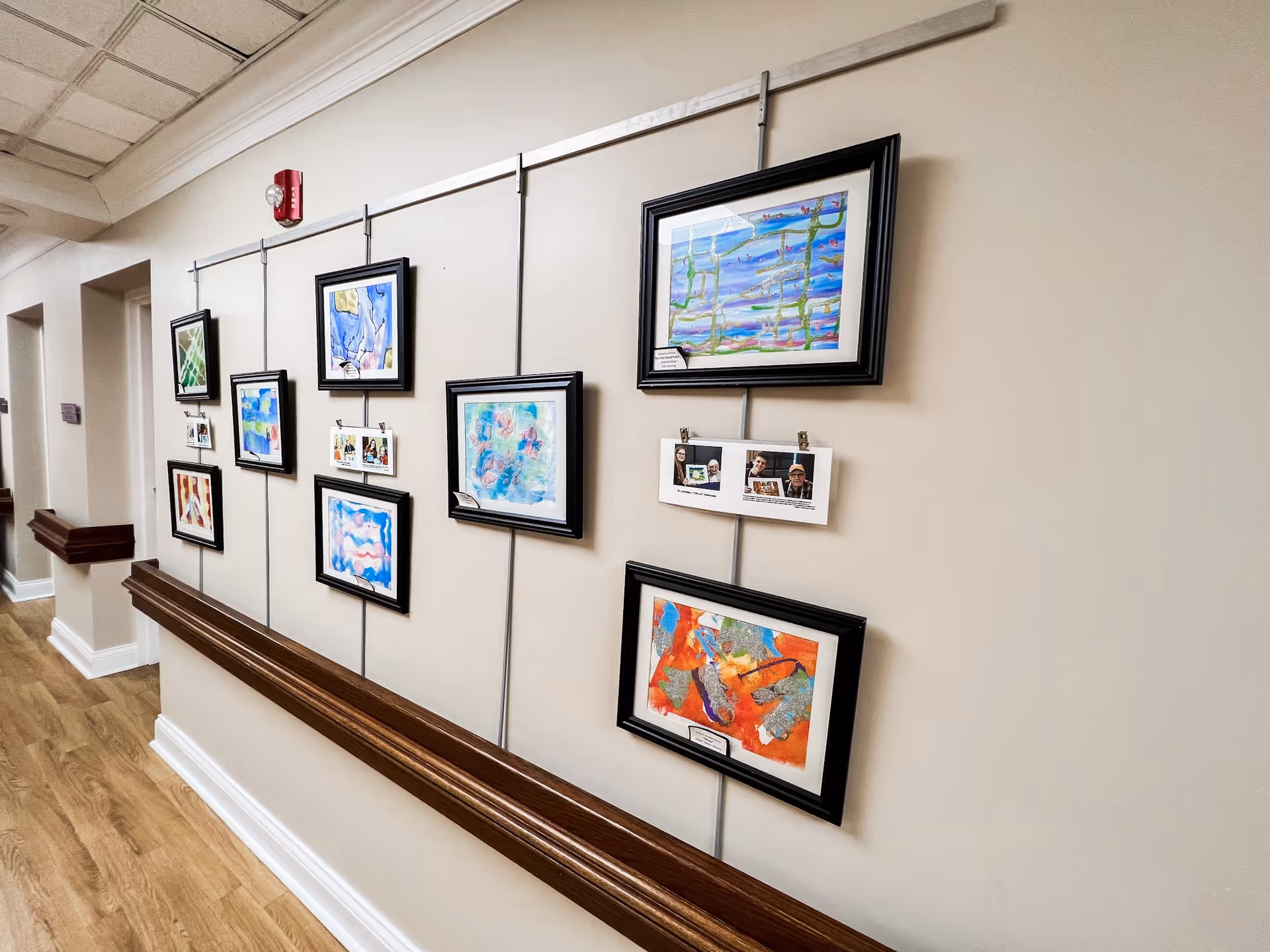 A hallway in a senior living facility with several framed colorful paintings and photographs displayed on a beige wall. The floor is wooden, and there are wooden handrails along the wall. The ceiling has white tiles and a fire alarm is visible near the top of the wall.