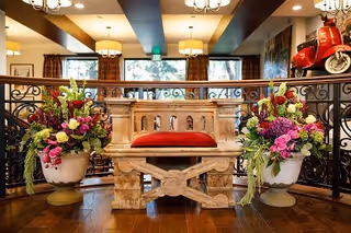 Ornate wooden bench with a red cushion flanked by two large floral arrangements in a warmly lit interior common area.