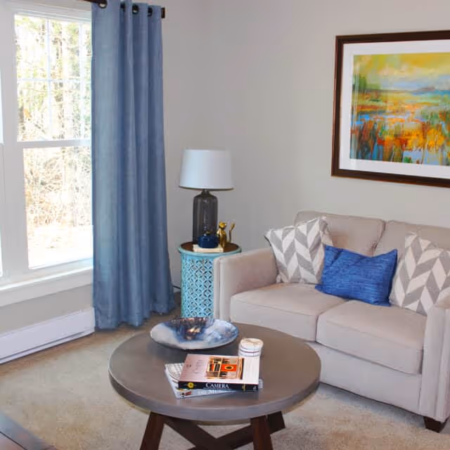 A cozy living room with a beige sofa adorned with two gray and white patterned pillows and one blue pillow. A round wooden coffee table in front of the sofa holds a decorative bowl, a candle, and some books. To the left, there is a window with blue curtains, and next to the sofa is a small round side table with a lamp and decorative items. A colorful framed painting hangs on the wall above the sofa.