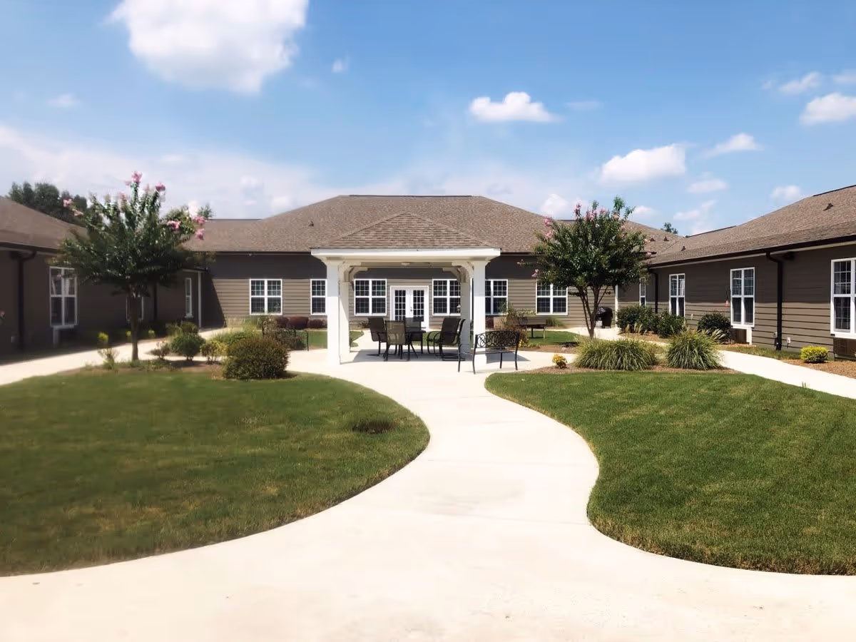 Outdoor courtyard area at Orchard Ridge Residences featuring a paved walkway, green lawns, small trees, shrubs, and a white gazebo with outdoor seating including chairs and a table under a clear blue sky.