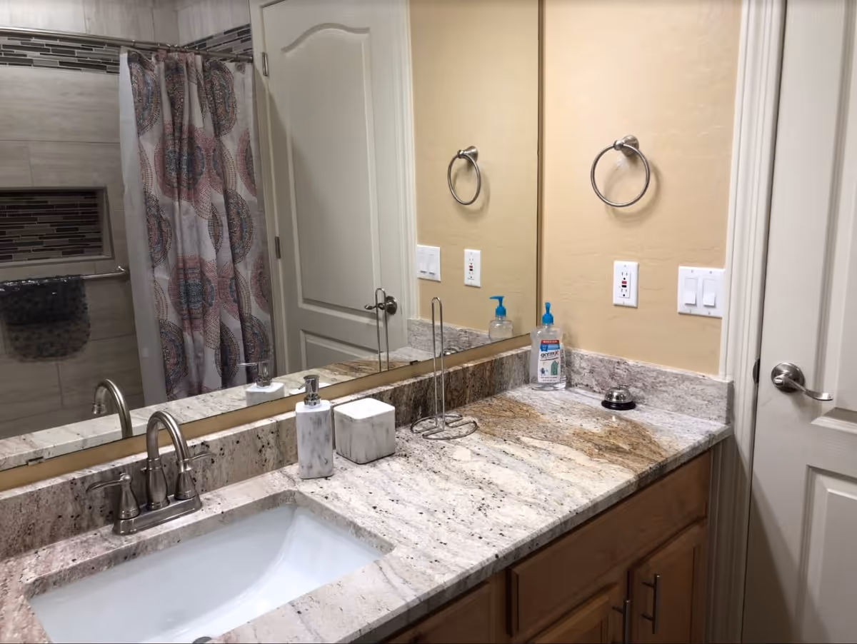A bathroom countertop with a sink, soap dispenser, tissue box, and hand sanitizer. A large mirror is mounted above the countertop. In the background, a shower area with a patterned shower curtain and tiled walls is visible. The bathroom has light-colored walls and wooden cabinetry below the countertop.