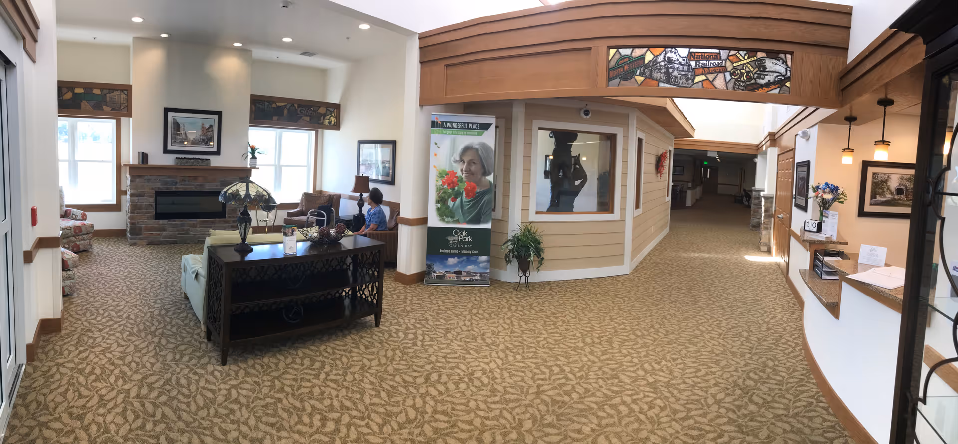 Interior view of a senior living facility lounge area with patterned carpet, a stone fireplace, and large windows. There are comfortable chairs and a sofa arranged around a coffee table with a decorative lamp. A person is seated on one of the chairs near the windows. To the right, there is a reception desk with flowers and framed pictures on the wall. A banner with an elderly woman holding flowers and the text 'Oak Park Place' is displayed near the center. The ceiling has wooden beams and decorative stained glass panels.