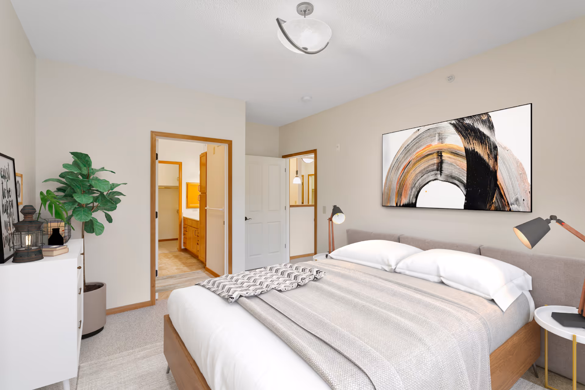 A bright bedroom with a large bed covered in neutral-colored bedding and two pillows. On the wall above the bed is a large abstract painting with black, orange, and gray brush strokes. There are two bedside lamps on either side of the bed. To the left, there is a white dresser with decorative items and a potted plant next to it. The room has beige walls and carpeted floors. An open doorway leads to a bathroom with wooden cabinetry and a walk-in closet visible.