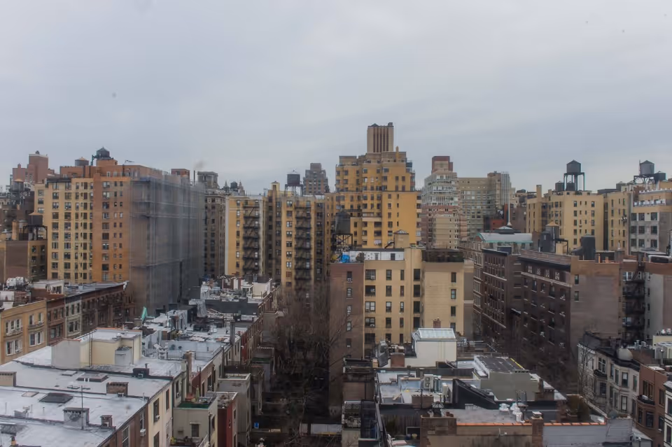 View of a cityscape featuring numerous mid-rise and high-rise buildings under a cloudy sky. The buildings vary in color and architectural style, with some having water towers on their rooftops.