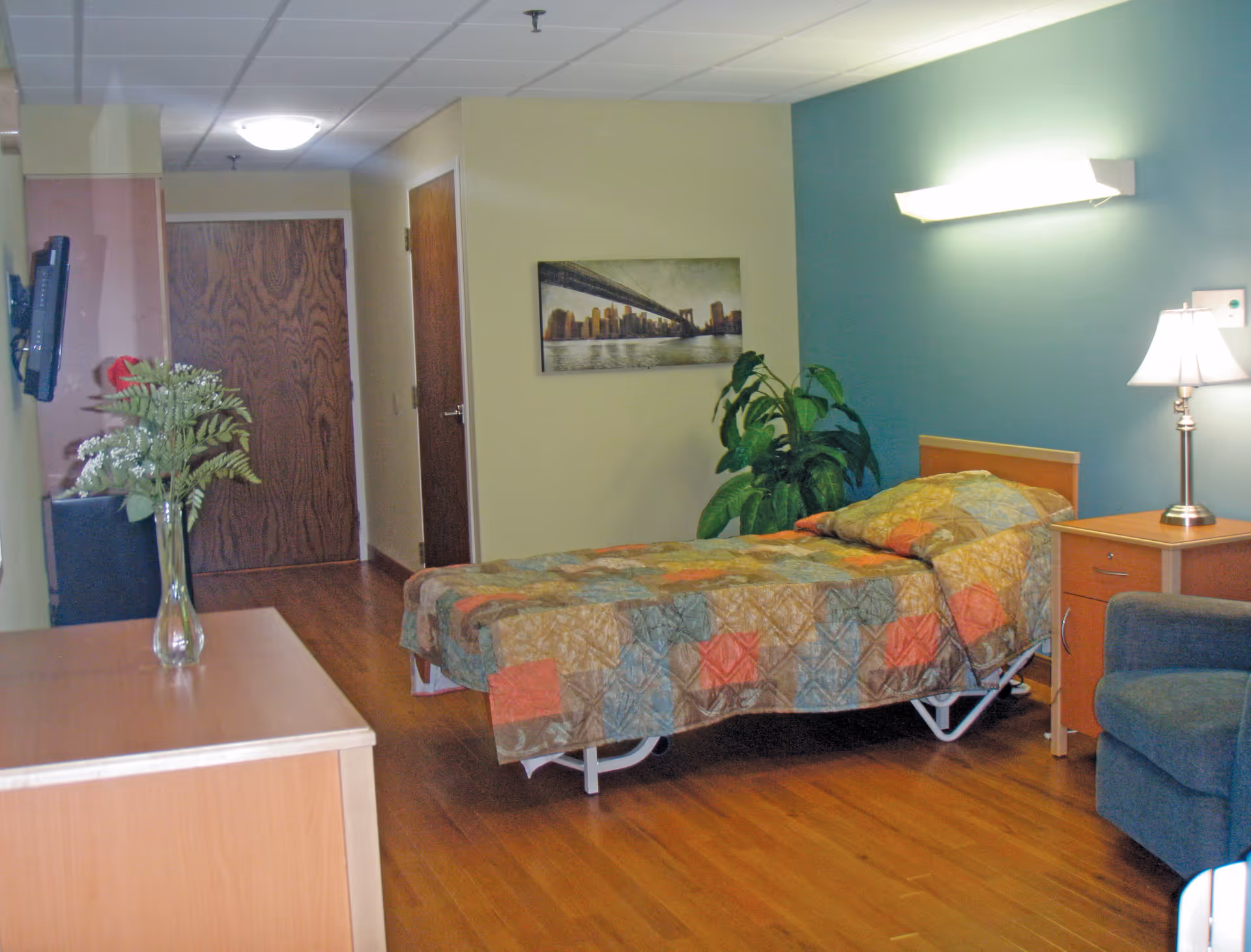 A single bed with a colorful patchwork quilt in a senior living facility room. The room has wooden flooring, a small wooden nightstand with a lamp, a green armchair, a potted plant, and a wall-mounted light fixture. There is a wooden door and a hallway in the background, with a flat-screen TV mounted on the wall and a vase with flowers on a wooden dresser.